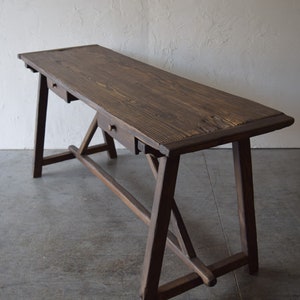 May include: A long, rectangular wooden desk with a dark brown stained finish and visible wood grain. The desk features two small drawers with round knobs, supported by an A-frame trestle base with a horizontal stretcher bar connecting the legs.
