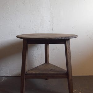 May include: A rustic, wooden side table with a round top and a triangular shelf. The table is made of dark-stained wood and has a simple, sturdy design. The table's height is approximately 24 inches.