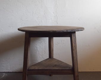 May include: A rustic, wooden side table with a round top and a triangular shelf. The table is made of dark-stained wood and has a simple, sturdy design. The table's height is approximately 24 inches.