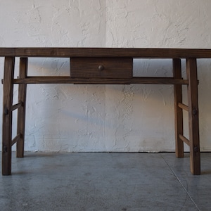 May include: A rustic wooden table with a single drawer and a simple design. The table is made of dark brown wood and has a natural finish. The legs are made of sturdy wood and are slightly angled.