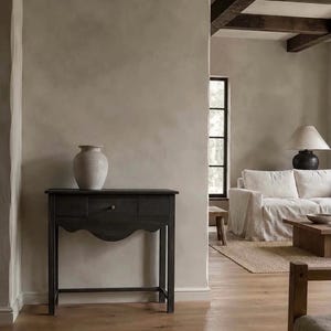 May include: A dark charcoal wooden console table with a single central drawer and a scalloped apron, featuring four straight legs. A matte light gray ceramic vase sits on the flat top surface of the table, which is positioned against a beige wall.