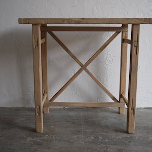 May include: A rustic wooden table with a simple, rectangular top and an X-shaped base. The table is made of natural wood with a light brown finish.