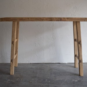 May include: A rustic wooden table with a long, narrow top and two sets of A-frame legs. The table is made of natural wood with a light brown finish and has a simple, minimalist design.