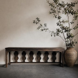 May include: A long, dark brown wooden console table with a flat rectangular top and a base featuring a series of vertical, scalloped wooden supports. Beside the table stands a large, textured, earth-toned clay vase containing a tall green plant.