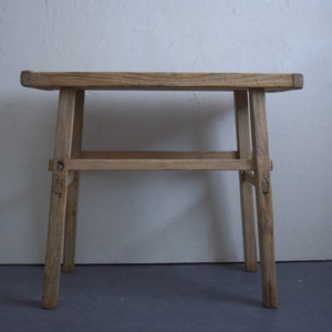 May include: A rustic, light-colored wooden stool with a rectangular seat and four angled legs. The stool has a simple design with visible wood grain and joinery details. It is approximately 18 inches tall and 12 inches wide.