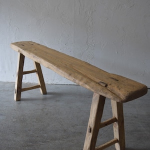 May include: A rustic wooden bench with a long, narrow top and two A-frame legs. The bench is made of natural wood with a weathered finish.