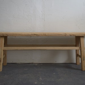 May include: A light-colored wooden bench featuring a long, rectangular seat with a visible natural grain. The bench is supported by four straight legs connected by horizontal stretchers on each side, showcasing a simple, functional construction.