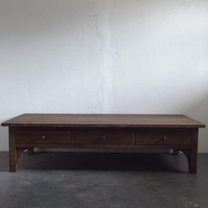 May include: A rectangular wooden coffee table with three drawers. The table is a warm brown color and has a simple, classic design. The drawers have round wooden knobs. The table is approximately 60 inches long.