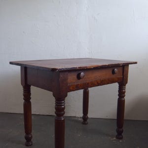 May include: A small, antique wooden table with a dark brown finish. The table has a rectangular top, a single drawer with two round knobs, and four turned legs. The table measures approximately 30 inches long, 20 inches wide, and 28 inches high.