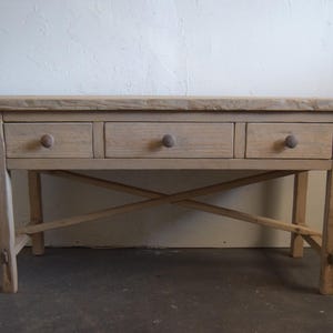 May include: A light wood console table with three drawers and a cross-shaped base. The table has a rustic, farmhouse style.