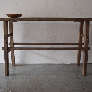 May include: A rustic wooden console table with a simple design. The table has a long, rectangular top and two sets of legs that are joined together by a crossbar. The table is made of unfinished wood and has a natural, weathered look.