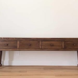 May include: A long, dark brown wooden console table with four drawers and rounded knobs. The table has four tapered legs with ball feet, and is set against a white wall and light wood floor.