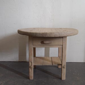 May include: A rustic wooden side table with a round top, a single drawer, and a lower shelf. The table is made of natural wood with a light brown finish.