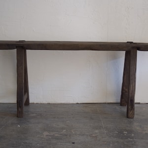 May include: A rustic wooden bench with a dark brown finish and a simple, sturdy design. The bench has a long, rectangular top and two pairs of angled legs.