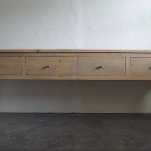 May include: A long, rectangular wooden console table featuring four horizontal drawers arranged in a single row. Each drawer is equipped with a small, round wooden knob. The table is supported by four tapered legs that terminate in rounded feet.