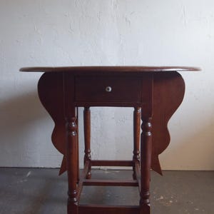 May include: A dark brown, antique drop-leaf table with a single drawer and a white knob. The table has turned legs and a decorative apron. The table is made of wood and is likely vintage.