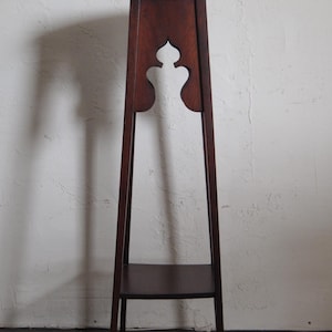 May include: A tall, dark wood plant stand with a round top and a lower shelf. The stand features a decorative cutout design and slender legs. The wood has a rich, warm tone, and the stand is set against a neutral background.