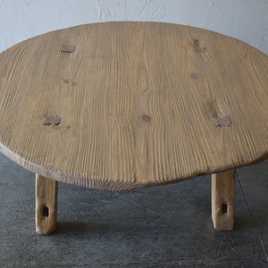 May include: A round wooden coffee table with a natural wood finish and four short legs. The table top has a slightly irregular shape and is made of a single piece of wood.
