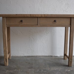 May include: A light brown wooden console table with two drawers and ring pulls. The table has a rectangular top and four slender legs. The wood has a natural, weathered appearance, and the table is set against a white wall.