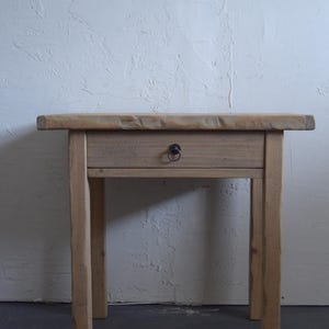 May include: A small, rustic wooden side table with a single drawer. The table is made of light-colored wood and features a dark metal ring pull on the drawer. The legs are square and sturdy. The table is approximately 24 inches tall.