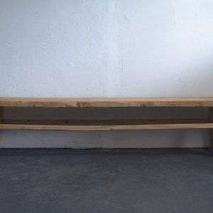 May include: A rustic wooden bench with a natural finish. The bench has a long, rectangular seat supported by angled legs and a lower stretcher. The wood grain is visible, and the bench appears to be handmade. The background is a textured white wall.