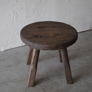 May include: A dark brown wooden stool with three legs and a round seat. The seat has a slightly uneven surface and is secured with metal nails.