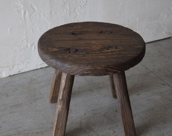 May include: A dark brown wooden stool with three legs and a round seat. The seat has a slightly uneven surface and is secured with metal nails.