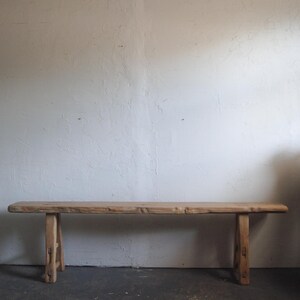 May include: A long, rectangular wooden bench featuring a natural light brown finish with visible wood grain. The bench has a flat, thick seat supported by four sturdy, angled legs connected by horizontal wooden crossbars on each side.