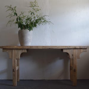 May include: A rustic wooden bench with a light finish, featuring a rectangular top and four sturdy legs. A large, textured vase filled with greenery sits atop the bench. The bench is approximately 60 inches long.