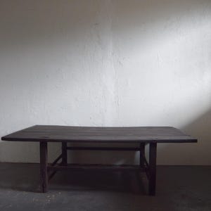 May include: A dark brown, rectangular wooden table with four legs. The table has a simple design and appears to be handmade. The table is set against a white wall and a dark gray floor.