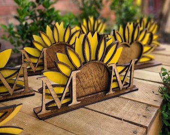 May include: Wooden napkin holders shaped like sunflowers with the word "MOM" in the center. The holders are painted yellow and brown.
