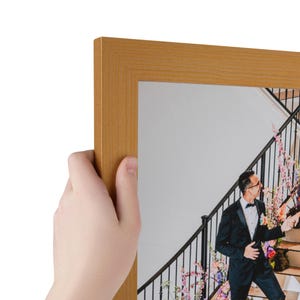 Puede incluir: Un marco de fotos rectangular con un acabado dorado mate y liso. El marco contiene una fotograf&iacute;a de una persona con esmoquin negro de pie en una escalera decorada con flores rosas y moradas, capturada en una impresi&oacute;n clara y n&iacute;tida.