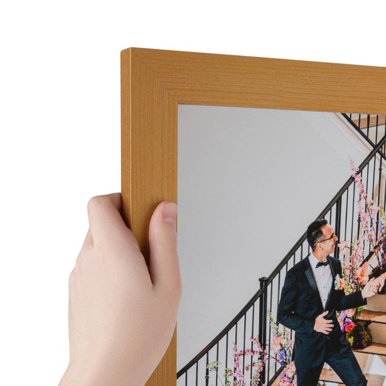 Puede incluir: Un marco de fotos rectangular con un acabado dorado mate y liso. El marco contiene una fotograf&iacute;a de una persona con esmoquin negro de pie en una escalera decorada con flores rosas y moradas, capturada en una impresi&oacute;n clara y n&iacute;tida.