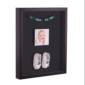 May include: A black shadow box frame with a baby photo, a pair of white baby shoes, and a string of colorful felt balls. The baby photo is of a smiling infant. The shoes have a bow on the front.