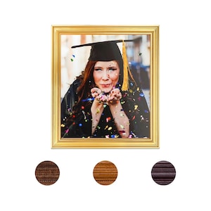 May include: A gold-framed picture of a woman in a graduation cap and gown blowing confetti. The woman is smiling and looking at the camera. The background is blurred and out of focus.