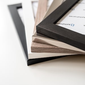 May include: A stack of rectangular picture frames in various finishes including matte black, solid white, light brown wood grain, and metallic silver. The top frame features a white insert with the blue text ArtToFrames printed on the surface.