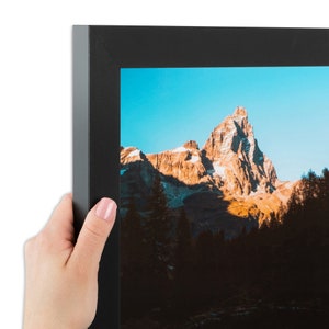 May include: A black picture frame with a landscape photograph of a mountain range with a blue sky and a bright sun. The mountain range is brown and has snow on the peaks. The frame is made of wood and has a simple design.