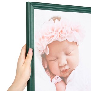 May include: A dark green picture frame containing a photograph of a sleeping infant wearing a light pink floral headband. The frame has a smooth, matte finish and a simple, clean-lined border design that surrounds the portrait of the baby.