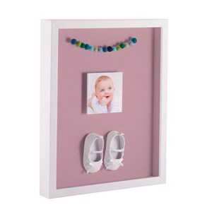May include: White picture frame with a pink background. The frame contains a photo of a baby, a garland of colorful pom-poms, and a pair of white baby shoes.