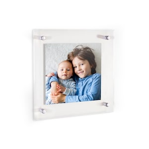 May include: Clear acrylic photo frame with a picture of two children. The photo is held in place by four silver-colored standoffs. The children are wearing blue shirts. The frame is square and has a white background.