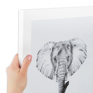 May include: A framed black and white pencil drawing of an elephant's face. The artwork is in a white frame, held by a hand. The elephant's detailed features are visible, including its large ears and trunk.