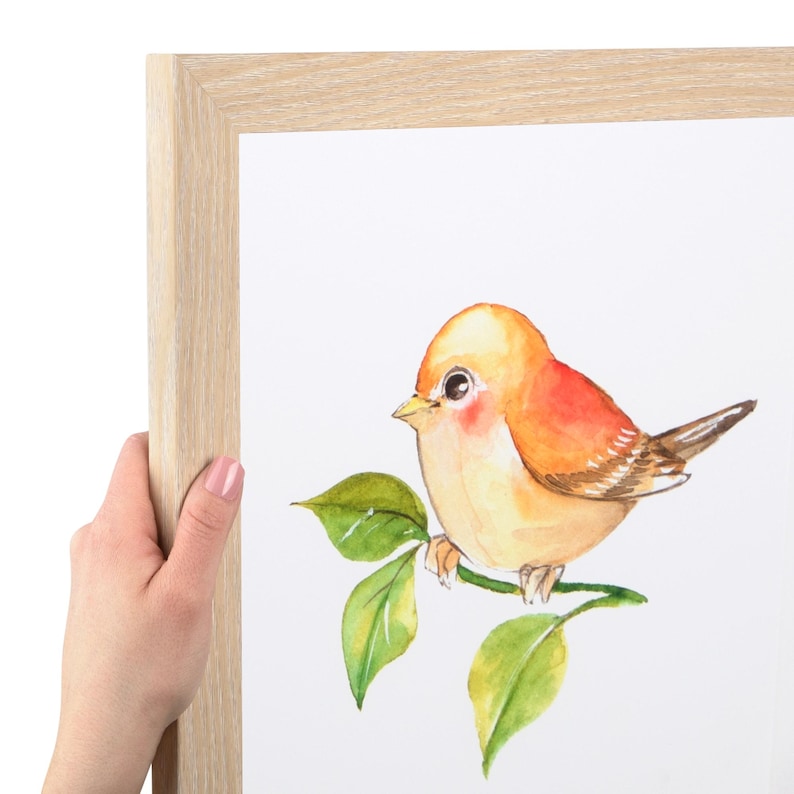 Puede incluir: Un marco de fotos de madera clara que contiene una impresi&oacute;n blanca de un p&aacute;jaro peque&ntilde;o y redondo con plumas naranjas y amarillas posado en una rama con hojas verdes. El p&aacute;jaro tiene ojos oscuros grandes y estilo de acuarela.