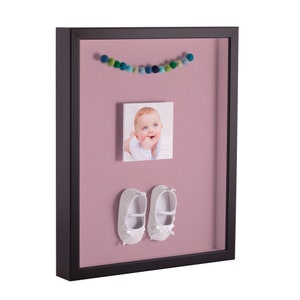 May include: A black picture frame with a pink mat. The frame contains a photo of a baby, a garland of colorful pom-poms, and a pair of white baby shoes.