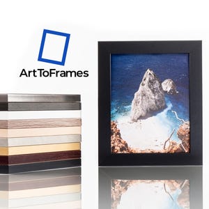 Puede incluir: Un marco de fotos negro que contiene una fotograf&iacute;a de un paisaje costero rocoso con agua azul, colocado junto a una pila vertical de muestras de marcos en diferentes colores y texturas, con el logotipo de ArtToFrames visible arriba.
