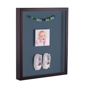 May include: A shadow box frame with a blue background, a photo of a baby, a garland of colorful pom-poms, and a pair of white baby shoes.