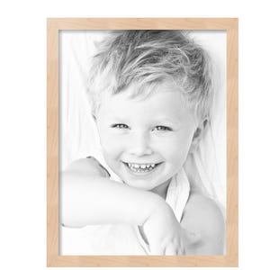 May include: A rectangular, light-colored wooden picture frame. The frame surrounds a black and white photograph of a smiling child with blonde hair. The child is wearing a white top.