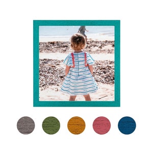 May include: A young girl in a blue and white striped dress with red and orange trim stands on a sandy beach, facing away from the camera. The girl is wearing a white dress with a blue and white striped pattern and red and orange trim. The background is a sandy beach with blue water in the distance.
