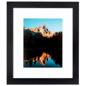 May include: A framed photograph of a mountain peak reflected in a still lake. The mountain is a light brown color with a dusting of snow on the top. The lake is a deep blue color and the sky is a light blue color.