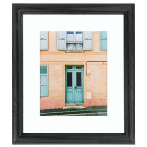 May include: A vintage-style photograph of a building with a teal door and windows with shutters. The building is painted a light peach color with a pink trim. The door has two panels and a small window in the top panel. The windows have white curtains and teal shutters.