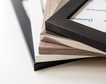 May include: A stack of picture frames in various colors, including black, white, and a light wood grain. The frames are made of wood and have a simple, modern design. The frames are stacked on top of each other.